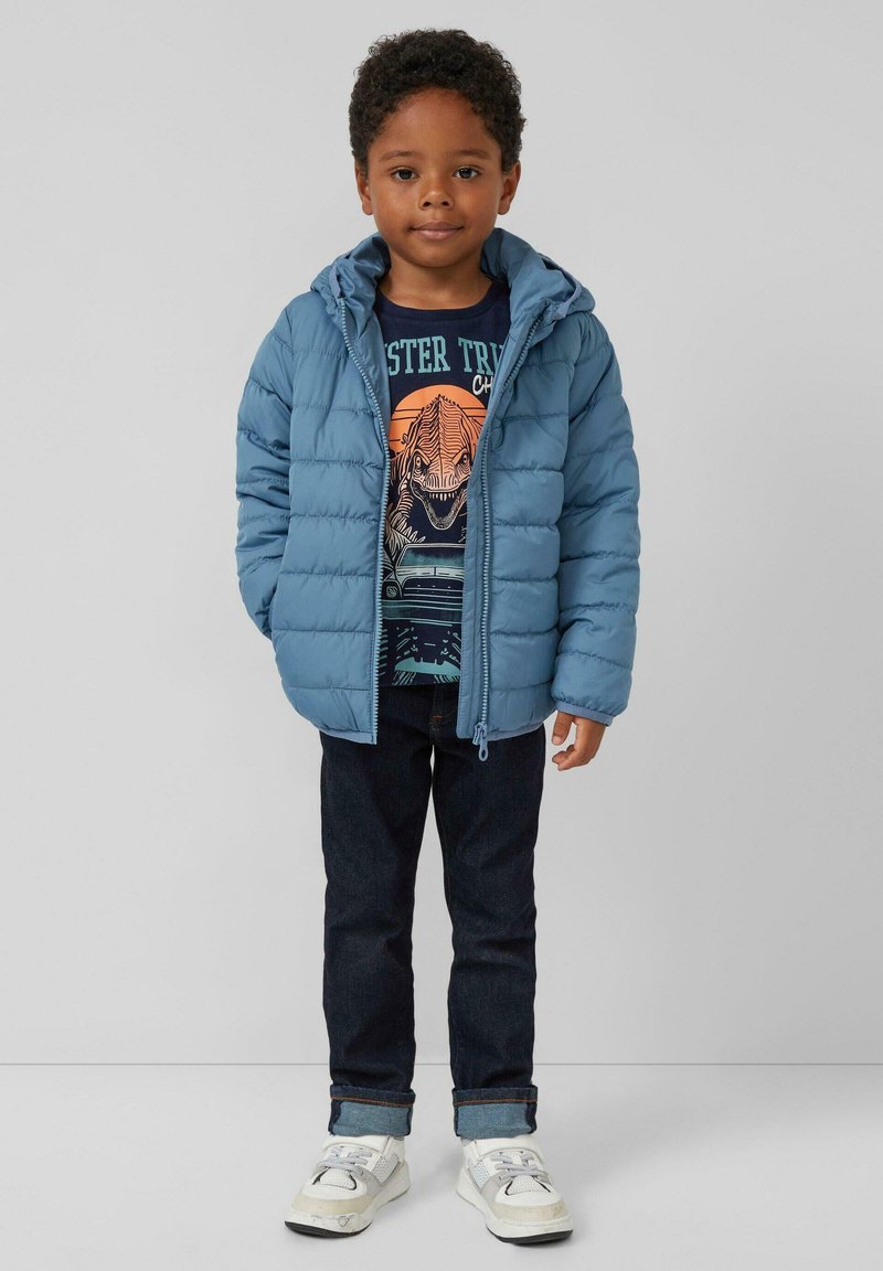 Lichtblauwe pufferjack met een capuchon, ritssluiting en horizontaal gestikt patroon, gedragen over een donker grafisch T-shirt en donkere jeans.