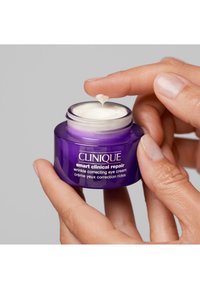 Clinique Smart Clinical Repair silmänhoitovoide purppurassa lasipurkissa, valkoisella voidepinnalla. Sormi levittää voidetta purkista.