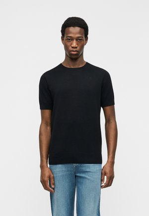 Jeune homme debout devant un fond blanc, portant un t-shirt noir uni à manches courtes et un jean bleu clair, face à la caméra.
