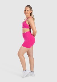 Roze sportbeha met een bandjesontwerp op de rug, gecombineerd met hoge roze shorts. Model draagt witte sneakers met zwarte accenten.