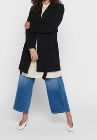 Žena v černém kabátu přes béžové šaty, širokých modrých džínách a bílých nízkých podpatcích stojí se založenýma rukama.