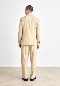Beige jakkesæt med en skræddersyet jakke og plisserede bukser. Har en spidse lapel, tre knapper og kontrasterende sorte knapper på ærmerne.