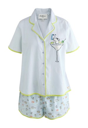 Vrouw staat binnen in een lichtblauw pyjamaset met cocktailglas en klein hondenmotief, ingelijste kunst aan de groene muur achter haar.