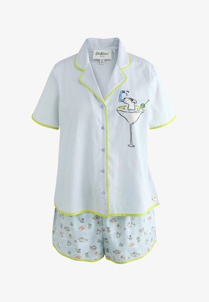 Vrouw staat binnen in een lichtblauw pyjamaset met cocktailglas en klein hondenmotief, ingelijste kunst aan de groene muur achter haar.