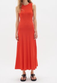 Červené bezrukávové šaty s žebrovanou strukturou, přiléhavým korzetem a skládací sukní. Modelka má na sobě černé sandály, čímž ukazuje délku šatů.