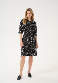 Musta mekko, jossa on valkoinen abstrakti kuvio, napitus edessä, kääntöhihoilla, vyötäröllä kiristettävä vyö ja polviin ulottuva suora leikkaus. Mustat kärkitossut.