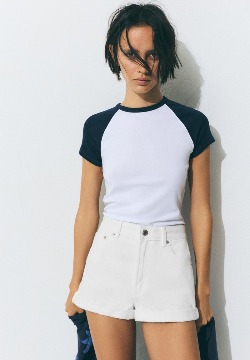 Jeune femme aux cheveux courts et foncés portant une chemise blanche à manches noires et un short blanc, debout devant un mur clair uni.