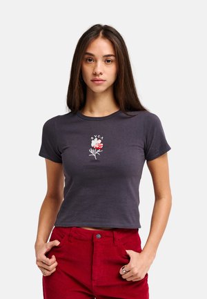 Jeune femme aux longs cheveux bruns portant un t-shirt court gris foncé avec un petit motif floral et un pantalon en velours côtelé rouge.