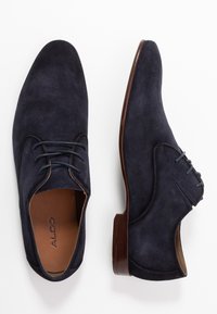 Chaussures habillées en suede bleu marine avec un bout arrondi, un laçage et un talon bas brun. Caractéristiques de surpiqûres sur les côtés.