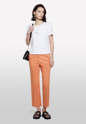 T-shirt en coton blanc avec un design graphique, associé à un pantalon orange à jambes droites. Sandales noires à semelle épaisse avec des accents en argent.