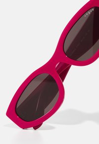 Ochelari de soare fuchsia cu rame în formă de lentilă și lentile întunecate. Prezintă un finisaj lucios și un detaliu subtil al logo-ului pe lateral.