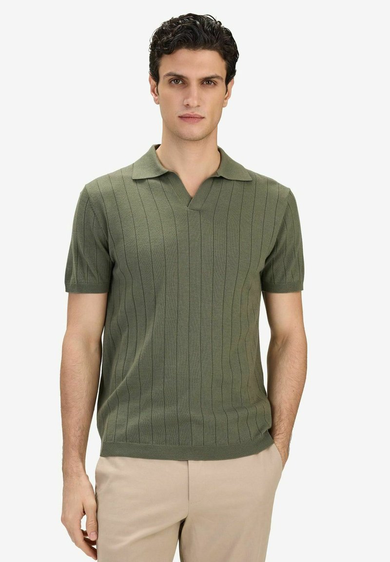 Homme aux cheveux bouclés foncés portant un polo en maille vert à manches courtes et un pantalon beige, posant devant un fond blanc uni.
