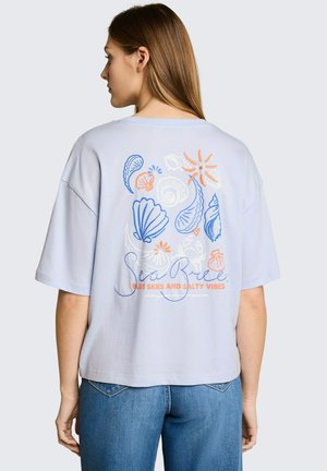Vrouw draagt een lichtblauw T-shirt met kleurrijke schelpen, zonontwerp en de tekst "Sea Breeze, Blue Skies and Salty Vibes" op de achterkant.