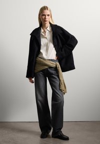 Cappotto nero texturizzato su una camicia bianca, con jeans a gamba larga scuri e una sciarpa beige annodata in vita. Modello che indossa scarpe nere.