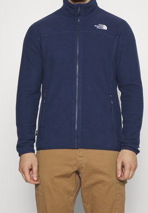 Mann trägt eine marineblaue Fleecejacke mit Reißverschluss und The North Face-Logo auf der Brust sowie beige Cargohosen.