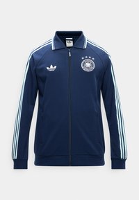 Navyblauwe sportjas met ritssluiting, lichtblauwe strepen op de mouwen, Adidas-logo op de linkerborst en embleem van de Duitse voetbalbond op de rechterborst.