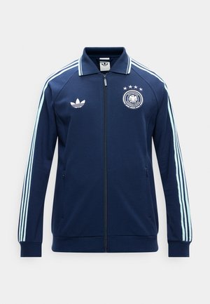 Chaqueta deportiva azul marino con cremallera, con rayas azul claro en las mangas, logo de Adidas en el pecho izquierdo y emblema de la Federación Alemana de Fútbol en el pecho derecho.