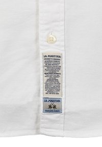 Bílá polokošile s knoflíkem, s modrými a šedými značkovými štítky obsahujícími text s podrobnostmi o původu produktu a jeho kvalitě.