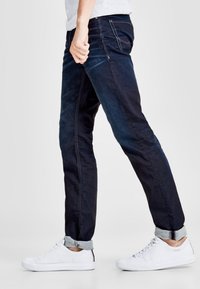 Jeans de mezclilla azul oscuro con un corte recto, que presentan una textura de lavado claro, puños doblados y bolsillos. Combinados con zapatillas blancas.