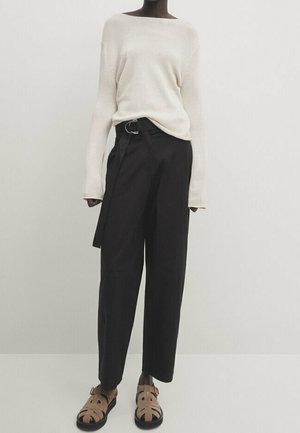 Personne portant un pull crème à manches longues, un pantalon large taille haute noir avec ceinture, et des sandales à boucle couleur camel, debout devant un fond uni.