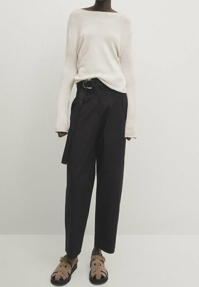 Personne portant un pull crème à manches longues, un pantalon large taille haute noir avec ceinture, et des sandales à boucle couleur camel, debout devant un fond uni.
