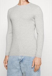 Maglione grigio chiaro a coste, maniche lunghe con scollo tondo e design aderente. Tessuto morbido con leggero effetto elasticizzato. Abbinato a jeans blu.