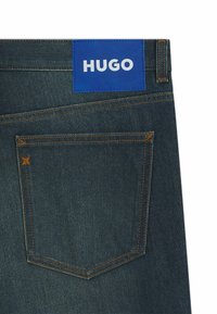Jeans in denim blu scuro con una prominente toppa in pelle blu etichettata "HUGO" in vita. Presenta cuciture arancioni e una tasca posteriore con una forma distintiva.
