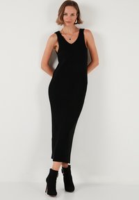 Black ribbed maxi dress with a v-neckline and sleeveless design. Features a fitted silhouette and side slits. Paired with black ankle boots.