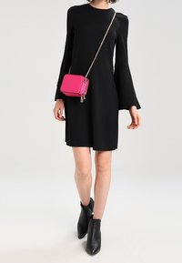 Robe noire à manches cloche avec un col arrondi, associée à un petit sac bandoulière rose et des bottines noires, créant un contraste saisissant.