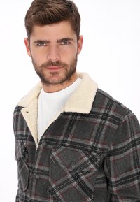 Chaqueta camisa de cuadros gris y negra con cuello de borrego sintético color crema, que cuenta con dos bolsillos frontales y cierres de botón.