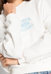 Bílá mikina s manžetovými rukávy a žebrovaným pasem. Obsahuje světle modrý vyšitý nápis "Los Angeles 1992." Měkká textura.