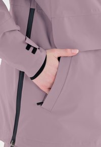 Vaaleanlila vedenpitävä takki, jossa on sivutasku ja musta yksityiskohta hihassa. Sujuva pinta ja tyylikäs muotoilu.