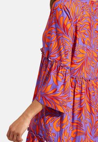 Jurk met een paarse achtergrond en oranje bloempatronen, met gerimpelde accenten op de mouwen en zoom, en een losse, vloeiende ontwerp.