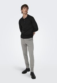 Schwarzes Langarmpolo mit drei Knöpfen, kombiniert mit hellgrauen, figurbetonten Hosen und schwarzen Stiefeletten. Schlichtes, reduziertes Design.