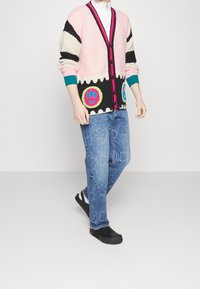 Meerkleurig gebreid vest met een ontwerp in roze, zwart en wit, voorzien van kleurrijke patchaccenten, gecombineerd met blauwe spijkerbroek met patroonafbeeldingen.