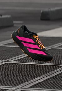 Fekete futócipő élénk rózsaszín csíkokkal és narancssárga fűzőkkel, fém rácsok felett lebegve egy városi utcán.