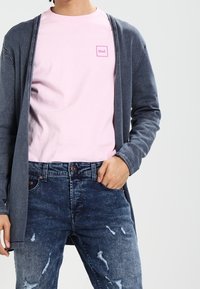 Graue Strickjacke mit weitem Schnitt über einem hellrosa T-Shirt mit kleinem Logo. Dunkelblaue Jeans mit Used-Look und sichtbaren Nahtdetails.