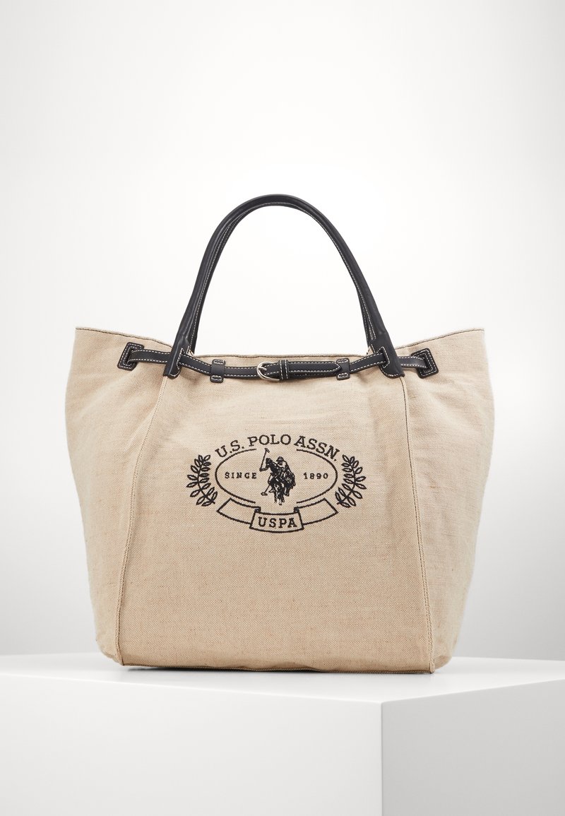 Beige canvas-handväska med svarta läderhandtag och detaljer. Den har en svart broderad logotyp och dekorativa sömmar.