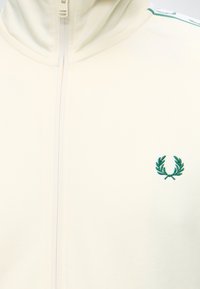Gros plan d'une veste crème zippée avec un logo couronne de laurier verte sur la poitrine gauche et des rayures assorties sur l'épaule.