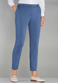 Pantalon bleu avec une coupe ajustée, présentant une texture lisse, des poches avant et une fermeture à boutons, associé à des mocassins de couleur claire.