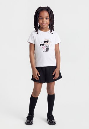 Νεαρή κοπέλα με πλεγμένα μαλλιά, που φοράει λευκό μπλουζάκι με καρτούν σχέδιο, μαύρη πλισέ φούστα, μαύρες κάλτσες ως το γόνατο και γυαλιστερά μαύρα παπούτσια.