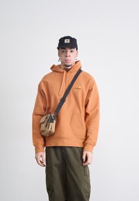 Orange bomulds-hoodie med frontlomme og logo, parret med olivenfarvede cargo-bukser. Båret med en sort kasket og en crossbody taske.
