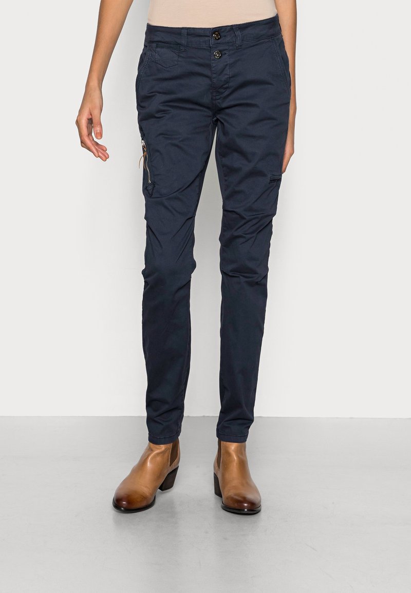 Person wearing navy blue slim cargo pants with side zipper pocket and tan leather ankle boots on gray floor.