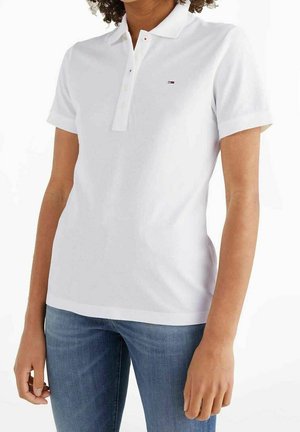 Kobieta w białej koszulce polo z krótkim rękawem i małym logo na piersi, ubrana w niebieskie dżinsy, stojąca na tle białego tła.