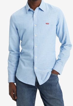 Mężczyzna w jasnoniebieskiej, długim rękawie koszuli zapinanej na guziki z małym czerwonym logo Levi's na piersi, w połączeniu z niebieskimi dżinsami.