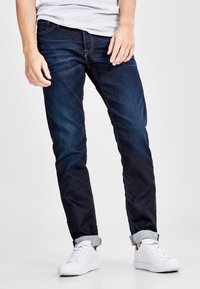 Pantalones vaqueros de denim azul oscuro con corte recto, que presentan detalles de costura sutil y puños doblados, combinados con zapatillas blancas.