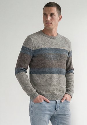 Mann trägt einen grauen Strickpullover mit blauen und braunen horizontalen Streifen, hellblaue Jeans und die Hände in den Taschen, schaut zur Seite.