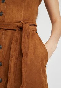 Braunes Wildlederkleid mit gesmokter Taille, Knopfleiste und Seitentaschen, das sich durch seine weiche Textur und glatte Oberfläche auszeichnet.