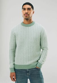 Hellgrüner gestrickter Pullover mit strukturiertem Muster, Rundhalsauschnitt und gerippten Bündchen und Saum. Getragen über blauen Jeans.