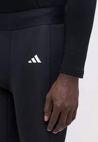 Schwarze sportliche Leggings mit glatter Textur, ausgestattet mit einem breiten Bund und dem weißen Adidas-Logo am Oberschenkel. Nahaufnahme einer Hand auf dem Stoff.